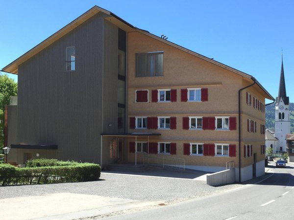 Der Adler von der Straße, Holzverschalung am Anbau, Rundschindeln und rote Läden am Altbau, dahinter der Kirchturm