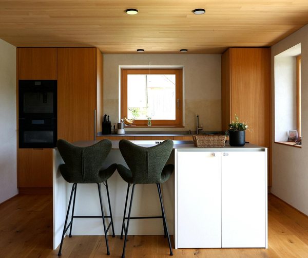 Die Küche mit Kochinsel und Holzfronten unter der Holzdecke, Fenster über der Arbeitsplatte